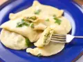 Pyszne pierogi ze szpinakiem i serem, udekorowane natką pietruszki, na niebieskim talerzu. Jedna porcja jest nadziana i widoczna na widelcu.