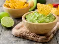 Pyszne guacamole w drewnianej misce, idealne do chrupania z nachos. Obok limonki i miska pełna chipsów.
