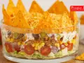 Warstwowa sałatka z nachosami w szklanej misie: sałata, pomidorki, cebula, kukurydza, mięso mielone, fasola, ogórek, śmietana i chrupiące nachos na wierzchu.