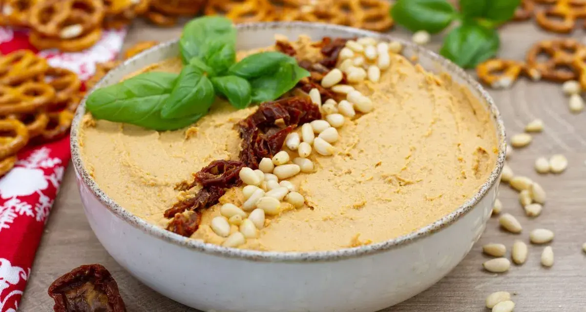 Aksamitny hummus z suszonymi pomidorami i orzeszkami pinii, udekorowany listkami bazylii. Idealny do chrupiących precelków.