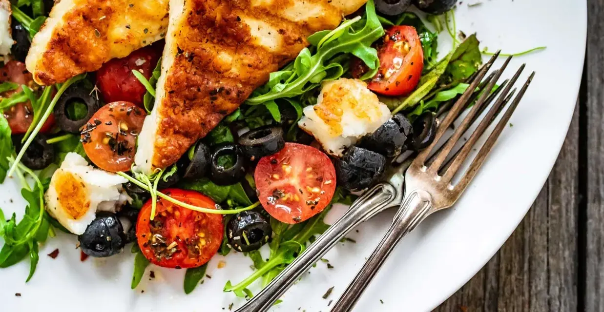 Pyszna sałatka z grillowanym halloumi, pomidorkami koktajlowymi i czarnymi oliwkami, podana na talerzu z widelcami.