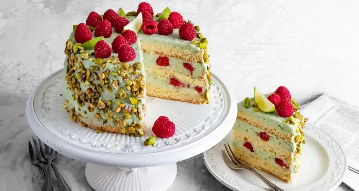 Wyjątkowy tort pistacjowo malinowy, udekorowany świeżymi malinami i pistacjami. Kawałek ciasta na talerzyku kusi warstwami biszkoptu i kremu.