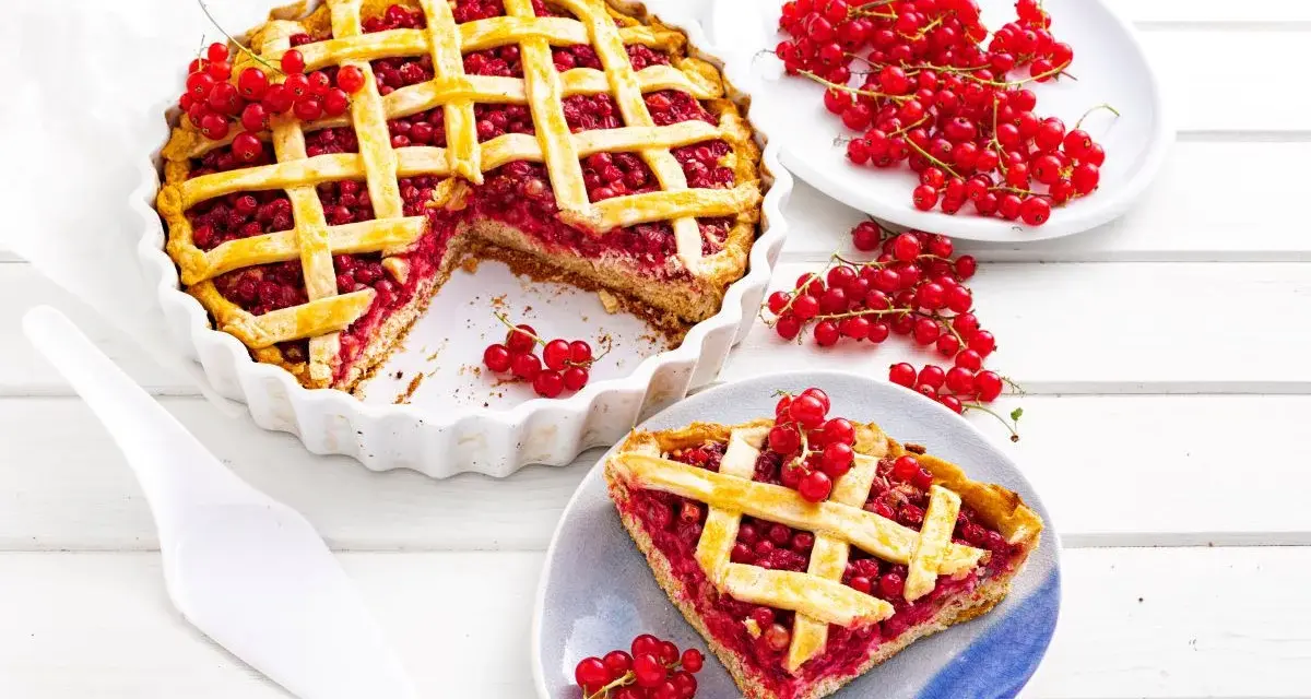 Pyszna tarta z porzeczkami, z kratką z ciasta i świeżymi owocami na wierzchu. Obok kawałek ciasta na talerzyku.