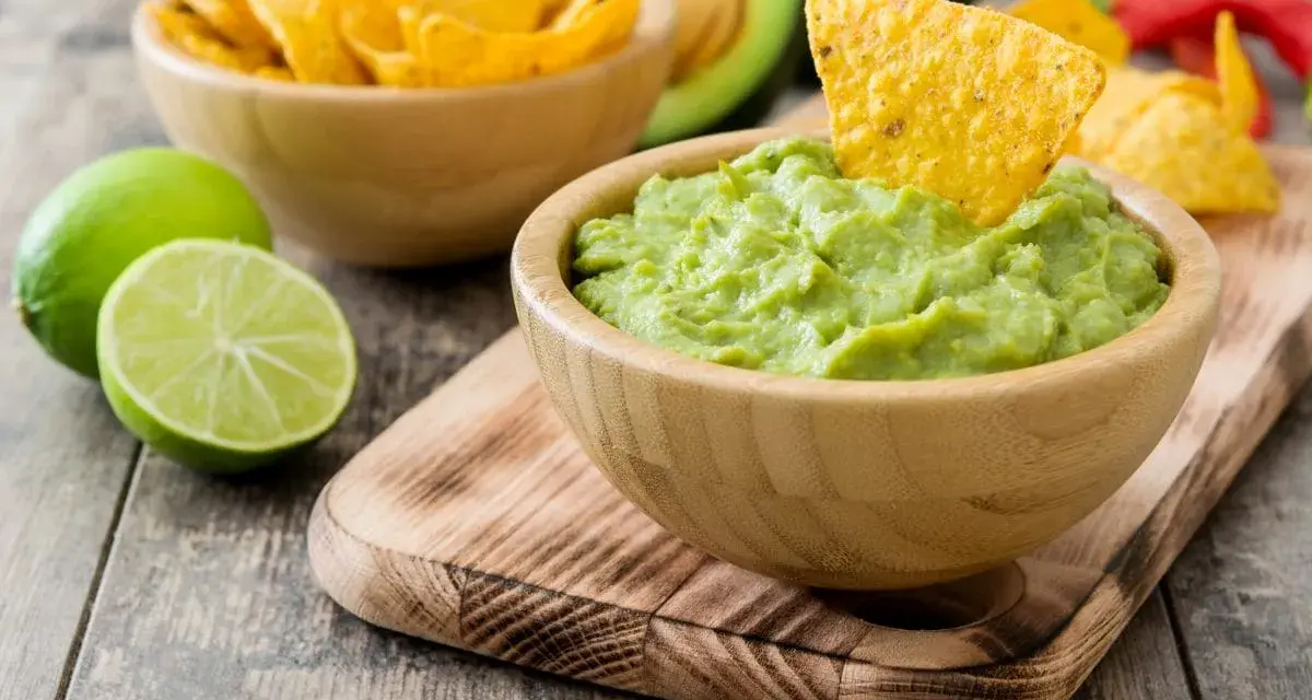 Pyszne guacamole w drewnianej misce, idealne do chrupania z nachos. Obok limonki i miska pełna chipsów.