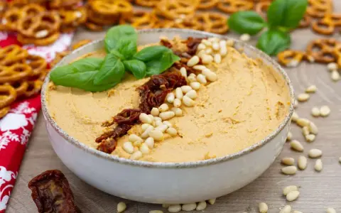 Aksamitny hummus z suszonymi pomidorami i orzeszkami pinii, udekorowany listkami bazylii. Idealny do chrupiących precelków.