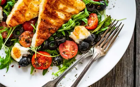 Pyszna sałatka z grillowanym halloumi, pomidorkami koktajlowymi i czarnymi oliwkami, podana na talerzu z widelcami.