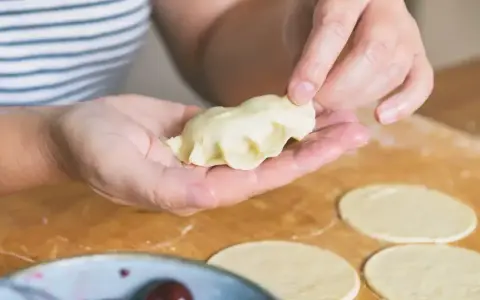 Ręce formują pierogi ruskie na drewnianej desce. W tle miska z wiśniami.