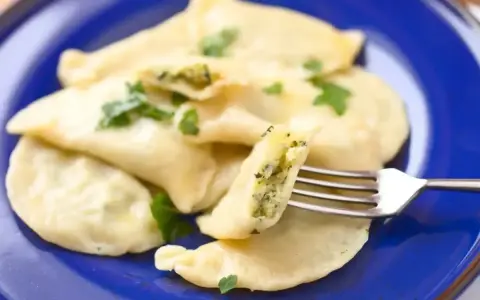 Pyszne pierogi ze szpinakiem i serem, udekorowane natką pietruszki, na niebieskim talerzu. Jedna porcja jest nadziana i widoczna na widelcu.
