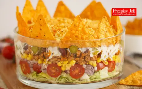Warstwowa sałatka z nachosami w szklanej misie: sałata, pomidorki, cebula, kukurydza, mięso mielone, fasola, ogórek, śmietana i chrupiące nachos na wierzchu.