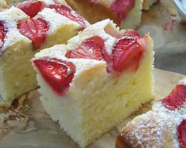 Puszyste ciasto na maślance z kawałkami truskawek, posypane cukrem pudrem. Idealne na letnie popołudnie.