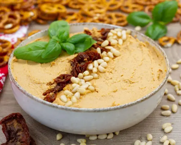 Aksamitny hummus z suszonymi pomidorami i orzeszkami pinii, udekorowany listkami bazylii. Idealny do chrupiących precelków.