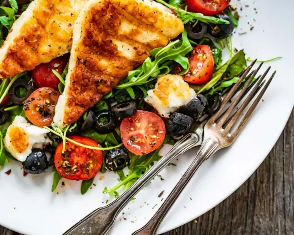 Pyszna sałatka z grillowanym halloumi, pomidorkami koktajlowymi i czarnymi oliwkami, podana na talerzu z widelcami.