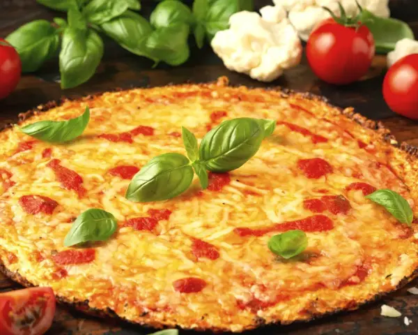 Pyszna pizza z kalafiora, z ciągnącym się serem i świeżą bazylią, otoczona pomidorami i różyczkami kalafiora.
