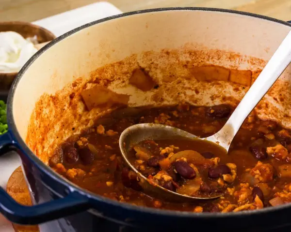 Gęste, aromatyczne chili con carne w garnku, z widoczną łyżką nabierającą porcję. Obok miseczka ze śmietaną.
