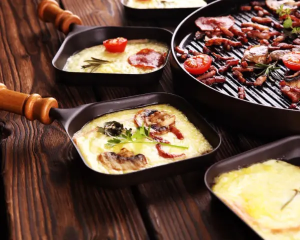 Raclette co to? Roztopiony ser z dodatkami w małych patelenkach i grillowane mięso z pomidorkami na drewnianym stole.