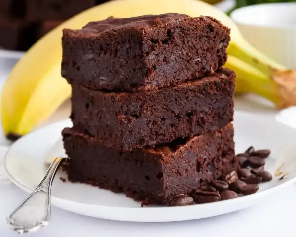 Stos puszystych brownie z bananami, ozdobiony ziarnami kawy, na talerzu z widocznym bananem w tle.
