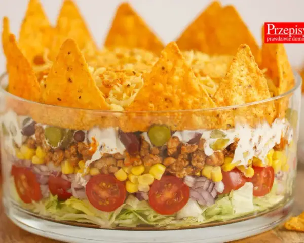 Warstwowa sałatka z nachosami w szklanej misie: sałata, pomidorki, cebula, kukurydza, mięso mielone, fasola, ogórek, śmietana i chrupiące nachos na wierzchu.