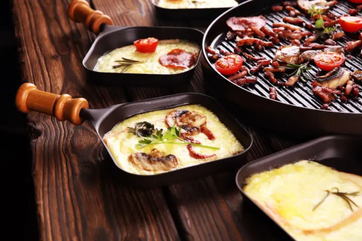 Raclette co to? Roztopiony ser z dodatkami w małych patelenkach i grillowane mięso z pomidorkami na drewnianym stole.