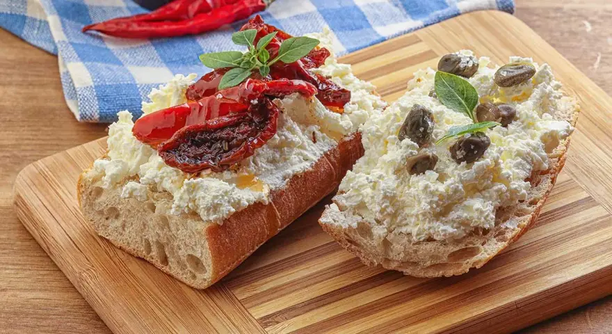 Dwa kawałki bagietki z twarogiem, suszonymi pomidorami i kaparami. Kapary dodają wyrazistego smaku, świetnie komponując się z serem i pomidorami.
