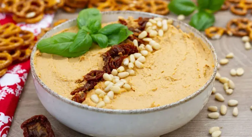 Aksamitny hummus z suszonymi pomidorami i orzeszkami pinii, udekorowany listkami bazylii. Idealny do chrupiących precelków.