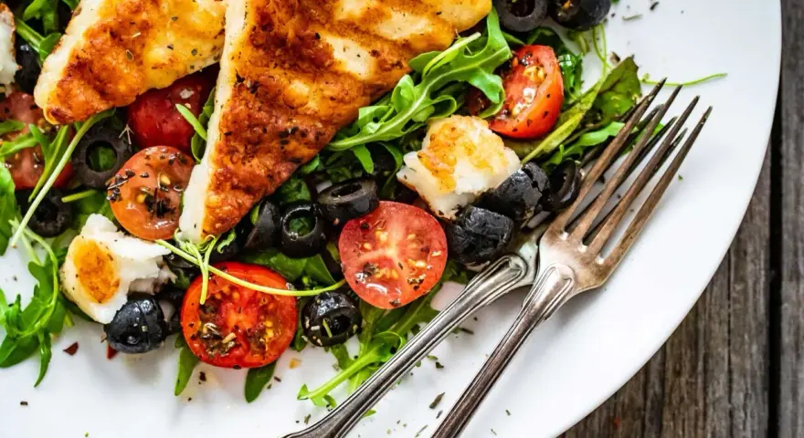 Pyszna sałatka z grillowanym halloumi, pomidorkami koktajlowymi i czarnymi oliwkami, podana na talerzu z widelcami.