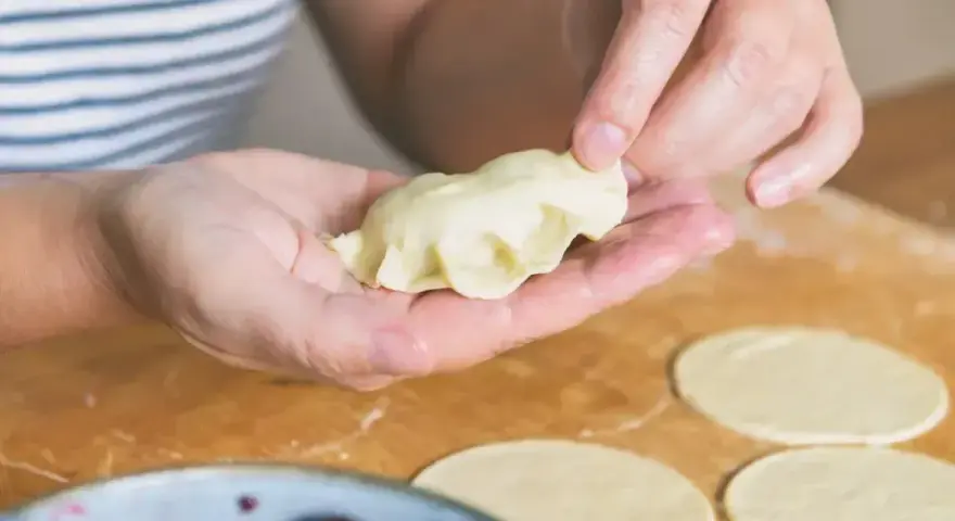 Ręce formują pierogi ruskie na drewnianej desce. W tle miska z wiśniami.