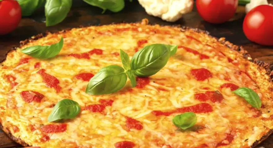 Pyszna pizza z kalafiora, z ciągnącym się serem i świeżą bazylią, otoczona pomidorami i różyczkami kalafiora.