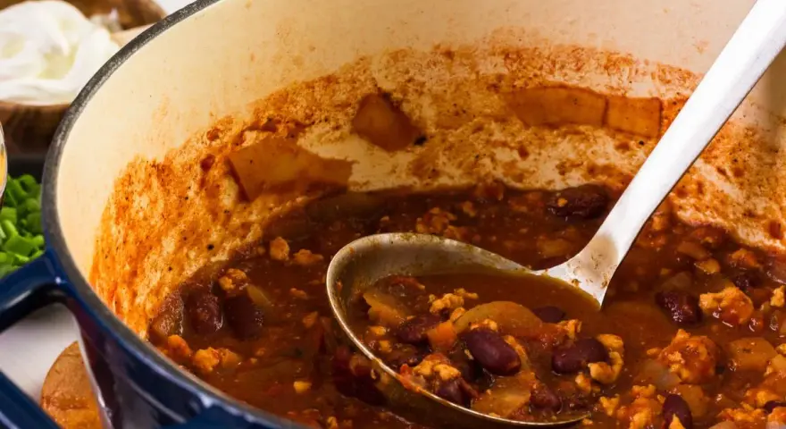 Gęste, aromatyczne chili con carne w garnku, z widoczną łyżką nabierającą porcję. Obok miseczka ze śmietaną.