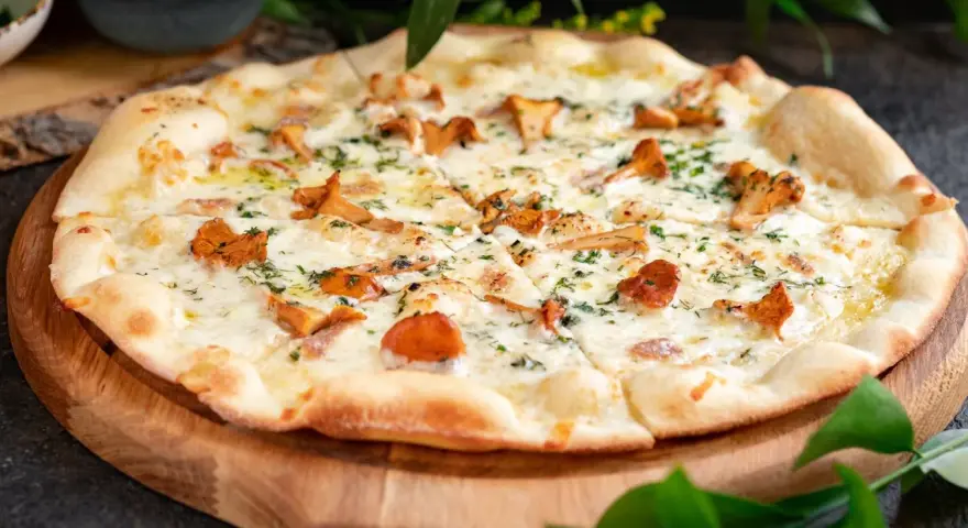 Pyszna pizza z kurkami, polana kremowym sosem i posypana świeżymi ziołami, podana na drewnianej desce.