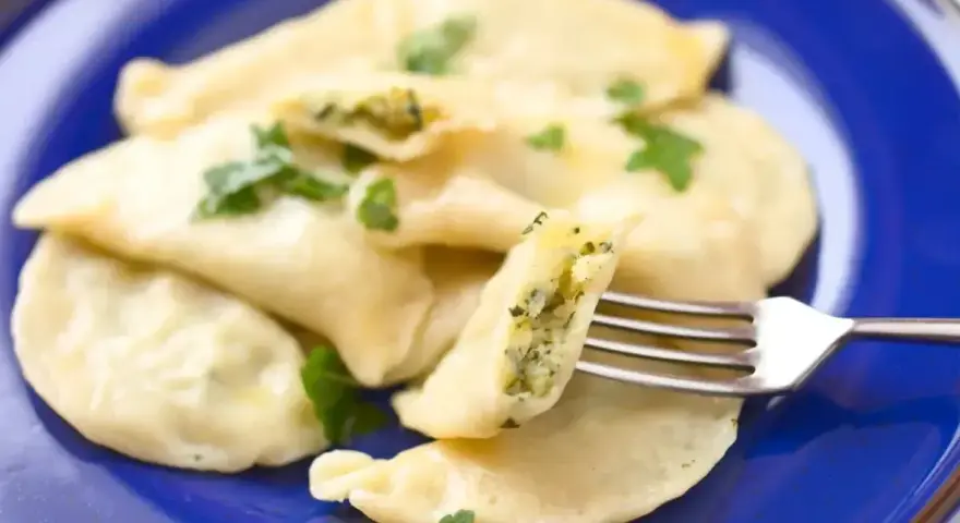 Pyszne pierogi ze szpinakiem i serem, udekorowane natką pietruszki, na niebieskim talerzu. Jedna porcja jest nadziana i widoczna na widelcu.