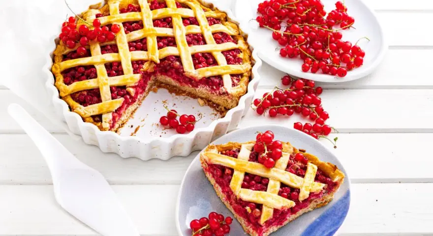 Pyszna tarta z porzeczkami, z kratką z ciasta i świeżymi owocami na wierzchu. Obok kawałek ciasta na talerzyku.