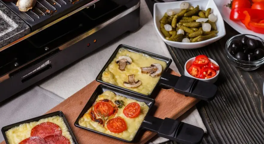 Raclette z pieczarkami i pomidorami, obok kiełbaski. Idealny ser do raclette rozpuszcza się w tych małych patelenkach.