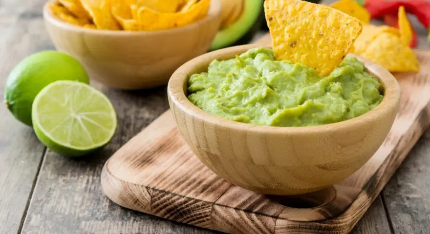 Pyszne guacamole w drewnianej misce, idealne do chrupania z nachos. Obok limonki i miska pełna chipsów.
