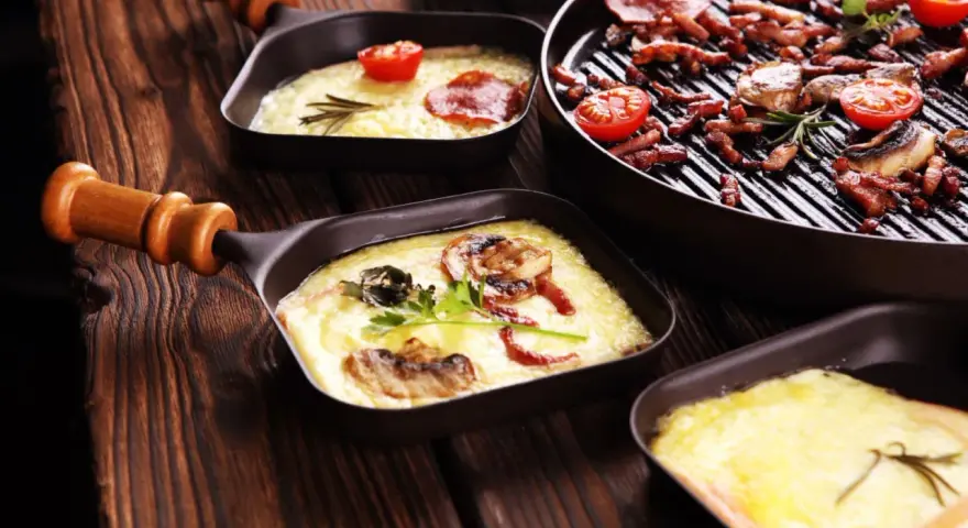 Raclette co to? Roztopiony ser z dodatkami w małych patelenkach i grillowane mięso z pomidorkami na drewnianym stole.
