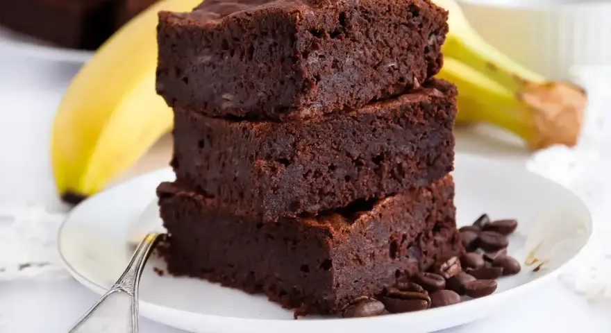 Stos puszystych brownie z bananami, ozdobiony ziarnami kawy, na talerzu z widocznym bananem w tle.