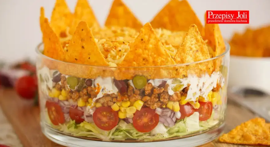 Warstwowa sałatka z nachosami w szklanej misie: sałata, pomidorki, cebula, kukurydza, mięso mielone, fasola, ogórek, śmietana i chrupiące nachos na wierzchu.