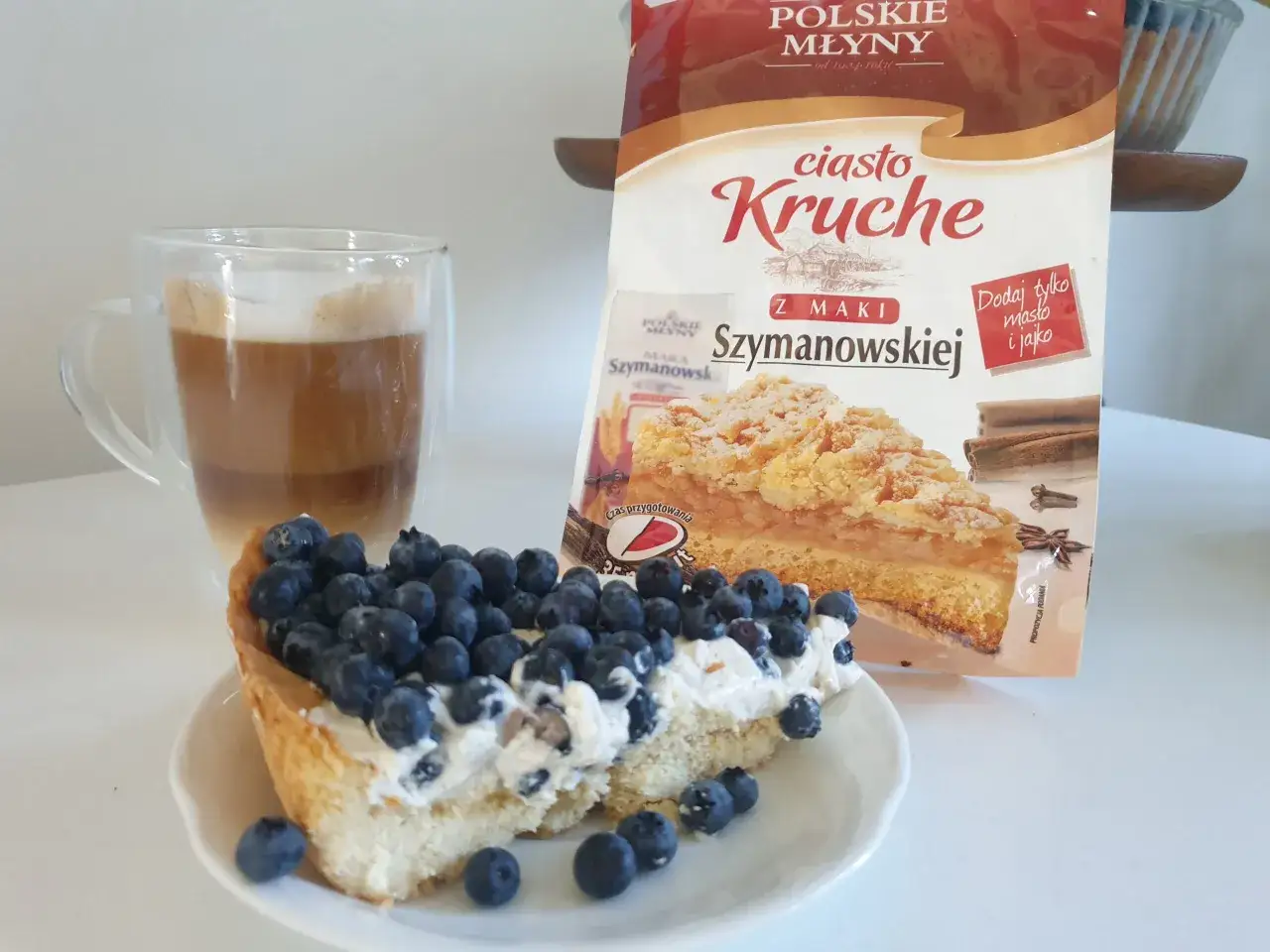 Pyszna tarta z borówkami i kremem, obok latte i opakowanie ciasta kruchego. Idealny deser.