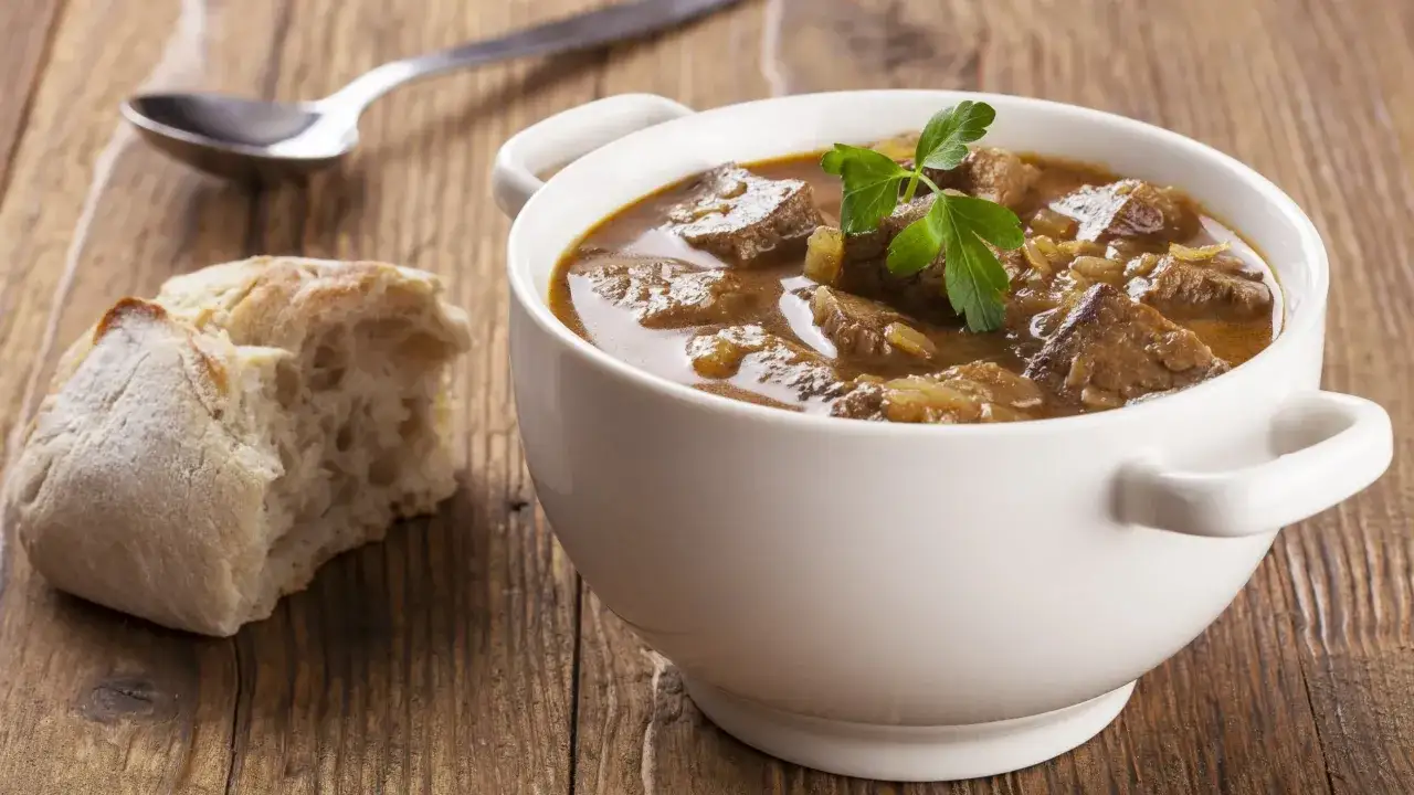 Gęsty, aromatyczny gulasz wołowy w białej miseczce, z gałązką pietruszki, obok chrupiąca bułka i łyżka. Idealny sposób, jak zagęścić gulasz.