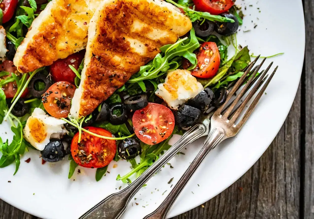 Pyszna sałatka z grillowanym halloumi, pomidorkami koktajlowymi i czarnymi oliwkami, podana na talerzu z widelcami.