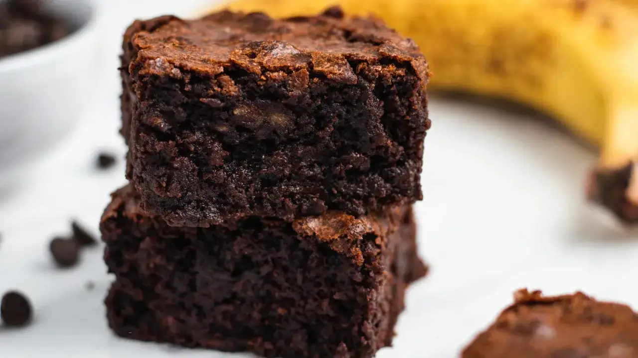 Dwa wilgotne brownie z bananami, ułożone jedno na drugim, z czekoladowymi kroplami i bananem w tle.