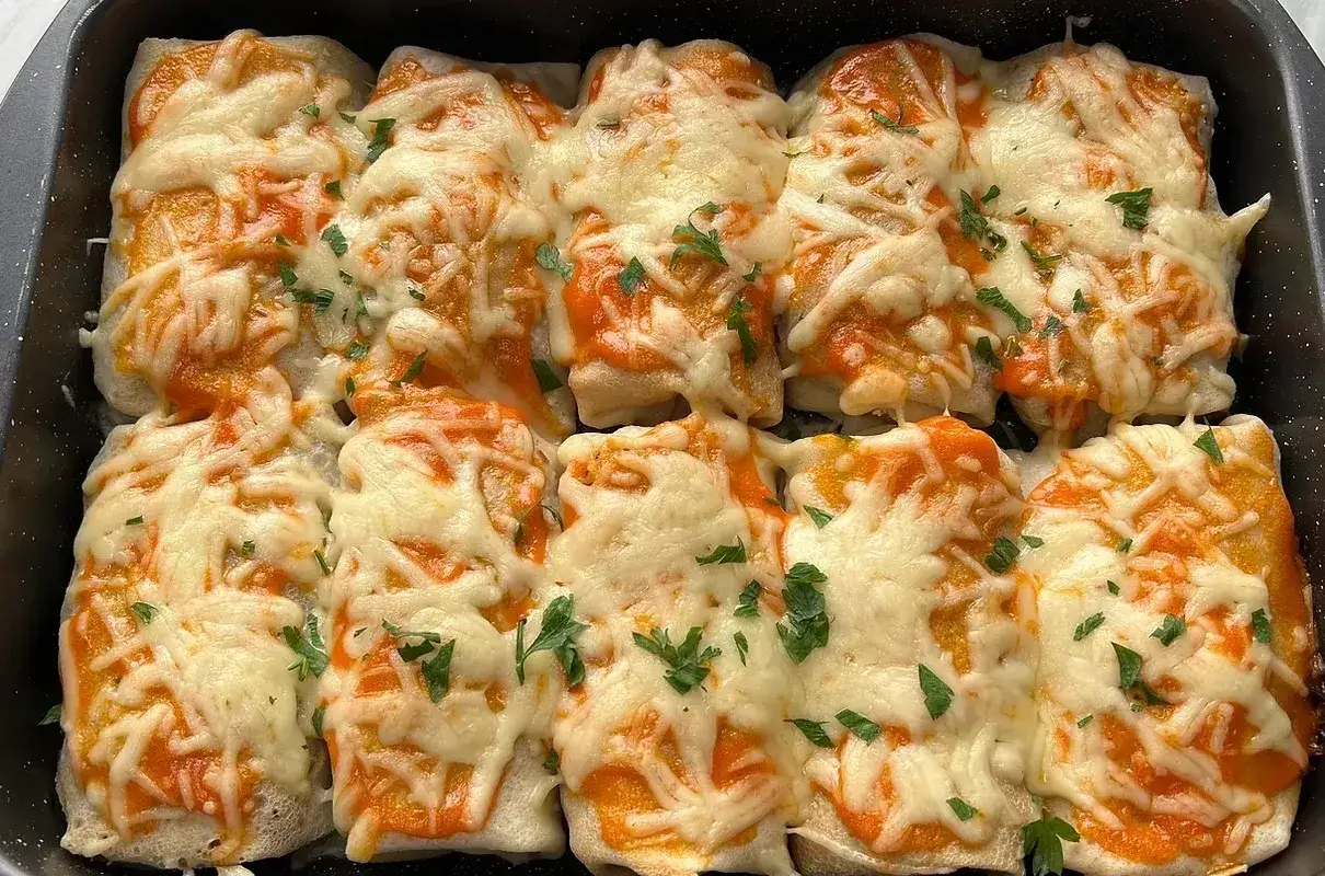 Zapiekane naleśniki z kurczakiem, polane sosem i posypane ciągnącym się serem, ozdobione natką pietruszki.