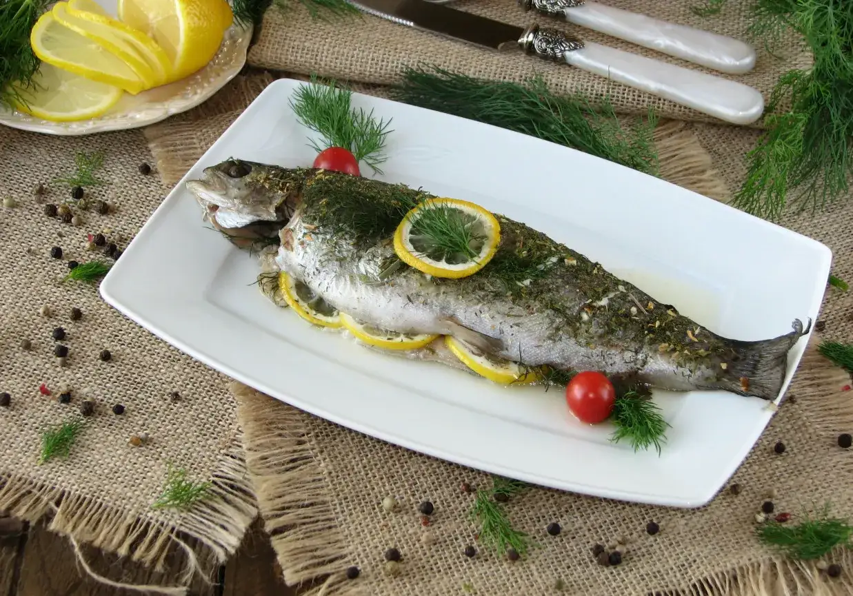 Pstrąg z piekarnika, pieczony z cytryną i ziołami, podany na białym talerzu, ozdobiony pomidorkami i koperkiem.