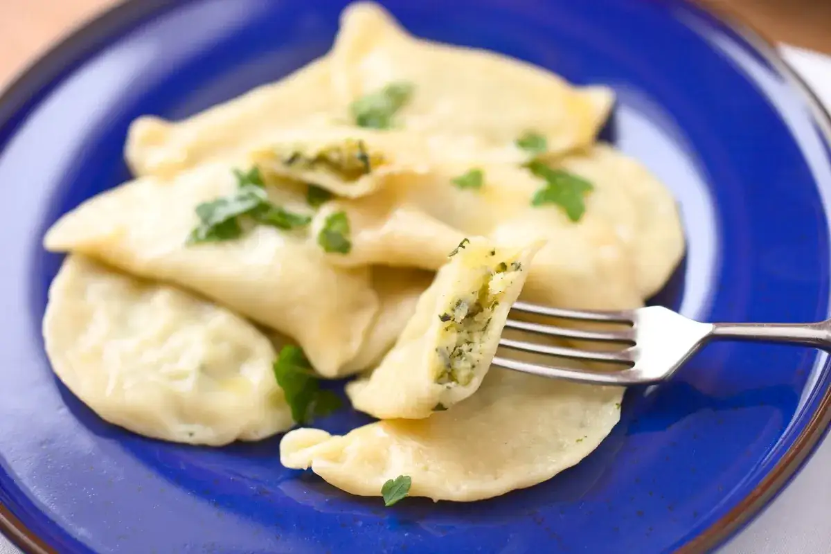 Pyszne pierogi ze szpinakiem i serem, udekorowane natką pietruszki, na niebieskim talerzu. Jedna pierożka jest nadziana widoczna.